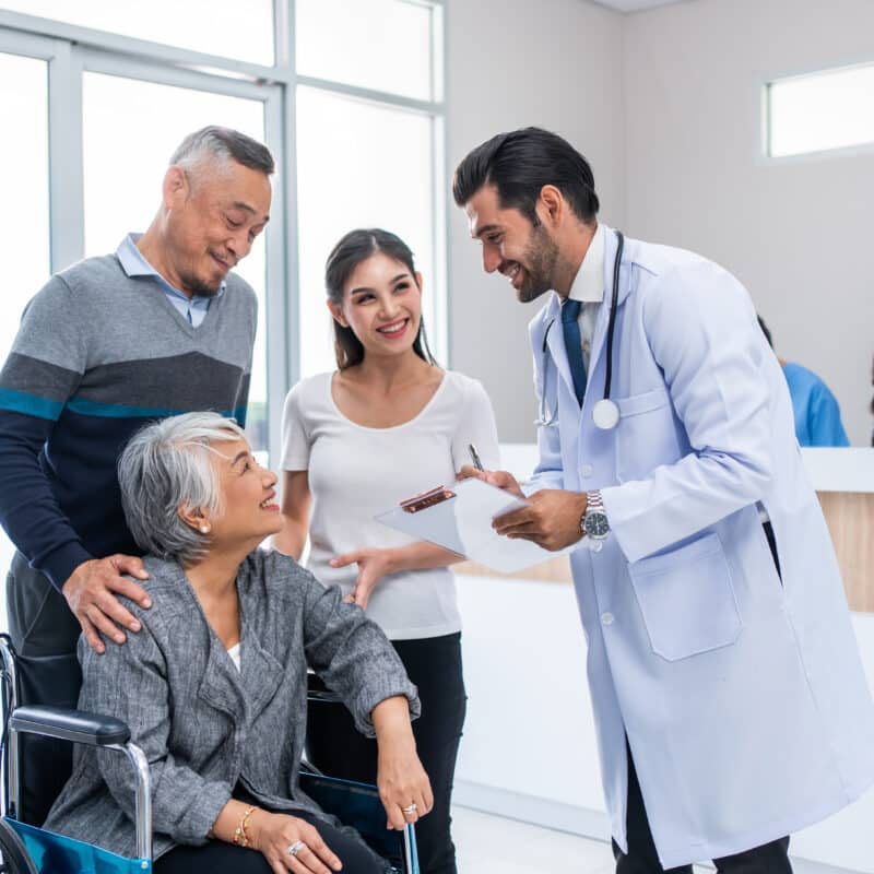 Un médecin souriant rassure une patiente âgée en fauteuil roulant et sa famille dans le hall d'une clinique.