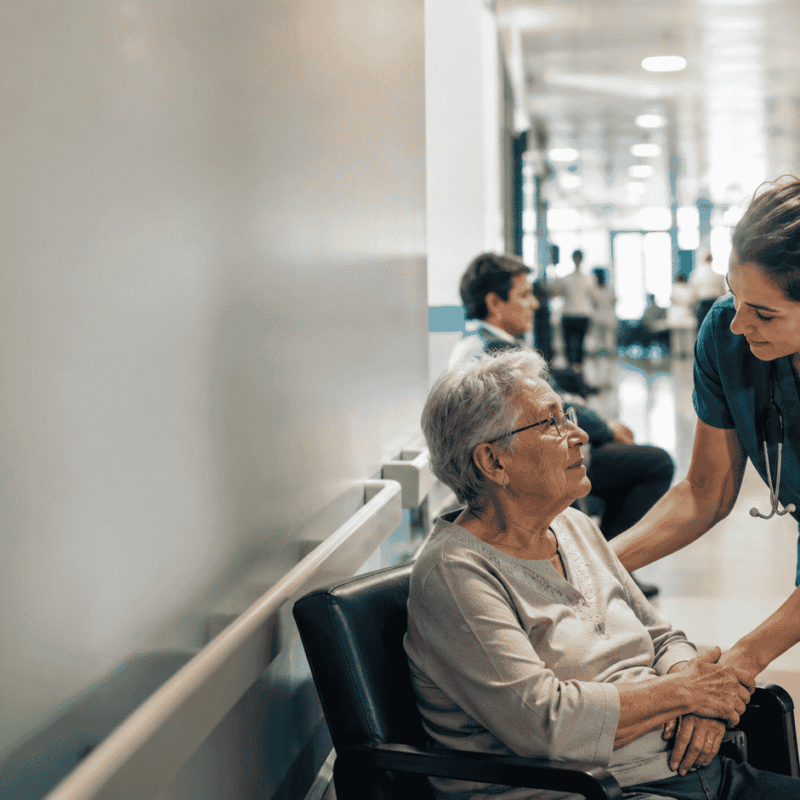 A calm, slightly wide view of a hospital corridor or emergency waiting area, an elderly patient being attended with care by a nurse, sense of a busy but humane hospital
